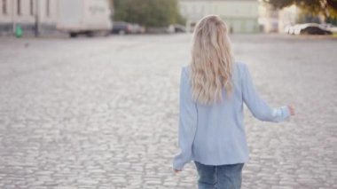 back view of a girl in headphones walking down the street dancing. a young blonde woman turns to the camera and smiles