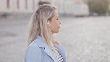 Close up portrait of stylish blonde young woman lady girl in headphones smiling at camera standing on city street in sunlight
