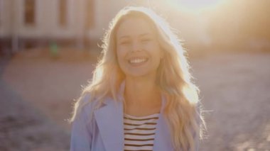 Beautiful fashionable model smiles, posing outdoors and straightens her long hair. Charming blonde  woman look in slow motion. Portrait of gorgeous girl in headphones looking to camera