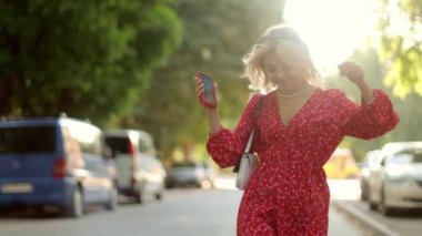Yazın şık elbiseler içinde mutlu bir sarışın kız sokakta yürüyor cep telefonundan müzik dinliyor, dans ediyor, dışarıda eğleniyor ve şarkı söylüyor.