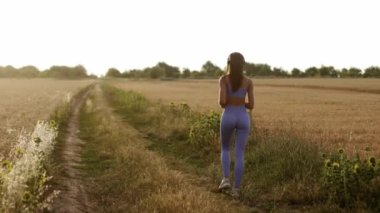 Camera follows slim young athletic woman jogging on field road outdoors at dawn in summer, concept of healthy running and outdoor exercise