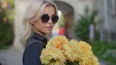 Close up portrait of young pretty stylish smiling blonde woman in sunglasses holding bouquet of flowers outdoors