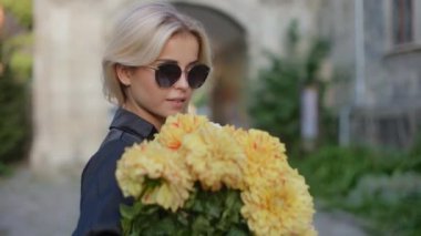 Close-up portrait of a young pretty stylish smiling blonde girl model in sunglasses posing for the camera and sniffing a bouquet of autumn flowers