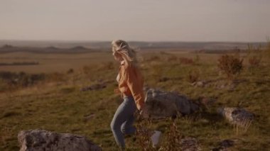 Slim girl woman blonde jumps on a big stone on a hill and raises her hands up in satisfaction, concept of freedom travel achievement success