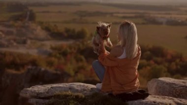  Young female girl sitting on the rock of a mountain peak at sunset, holding and kissing and hugging her little dog on a windy day. happy with a beautiful view smiling at the camera