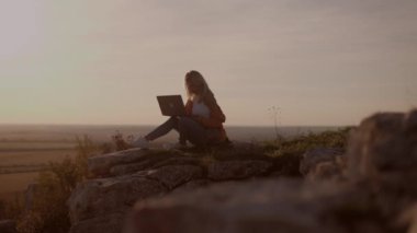 Side view, silhouette of a young woman sitting on a rock with an autumn landscape at sunset and closing a laptop after finishing work, holding a dog in her arms, working online, freelancer, student