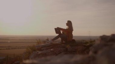 Side view, silhouette of a young woman sitting on a rock with an autumn landscape at sunset and closing a laptop after finishing work, holding a dog in her arms, working online, freelancer, student