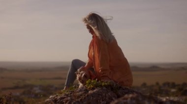 A young woman in an orange jacket sits on a mountain peak at sunset, holding her small dog in her arms on a windy day. happy watching the sunset with a beautiful view