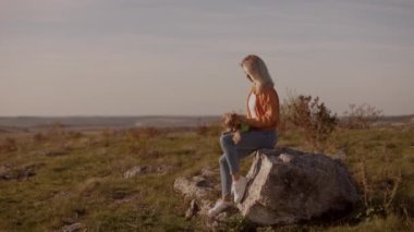 Güzel bir sonbahar manzarası olan bir tarlada taş üstünde oturan çekici sarışın kadın ve kollarında Yorkshire teriyeri köpeği, onu okşuyor ve kameraya gülümsüyor.
