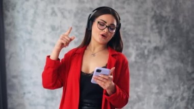 Positive European Caucasian young woman listening to music enjoying dancing with headphones on gray studio background. Radio, wireless modern sound technologies, smartphone player.