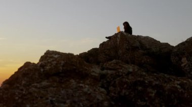 Woman working on laptop sitting on rock above river at sunset, beautiful view of yellow sky, female silhouette, outdoor work, relaxation, rest, alone, freelancer, work online.