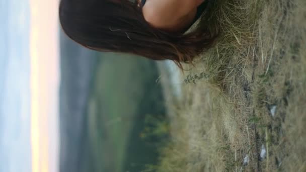 垂直方向のビデオオリエンテーション長い髪の少女を持つ若い女性は草の上に横になり 外で訓練した後 日没時に信じられないほどの景色で丘を見上げます — ストック動画