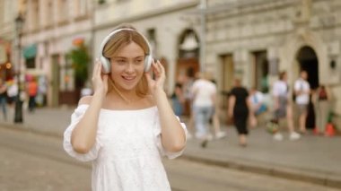 Genç, mutlu, sarışın, beyaz yaz elbisesi içinde şehir merkezinde yürüyen, müzik dinleyen bir kız. Kablosuz kulaklık takan mutlu genç bir kadının yavaş çekimi.