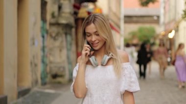 Genç, mutlu, güzel bir kadın cadde boyunca yürüyor, akıllı bir telefonla konuşuyor, gülümsüyor, hoş bir sohbet ediyor.