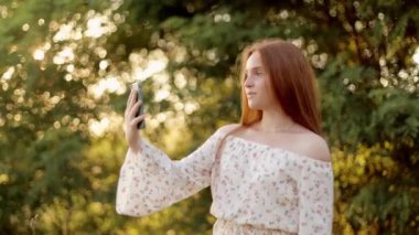 Güzel, mutlu, genç, boru saçlı kız bir video görüşmesi yapıyor, gülümsüyor, konuşuyor, yazın şık bir elbiseyle parkta rahatça duruyor, yakın plan..
