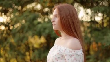 Güzel bir gün batımında parkta yürüyen kızıl saçlı çekici genç bir kız. Kendine yakın çekim. Model, kameraya bak, saçına dokun.