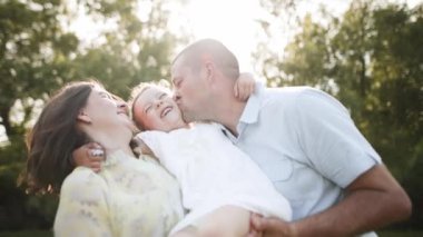 Mutlu aile, baba ve kızın yaz günbatımında parkta sarılmaları. Aile birlikte dışarıda tatilin tadını çıkarıyor. Ebeveynler çocuğu kollarına alır, kucaklar ve öperler. Babalık kavramı.