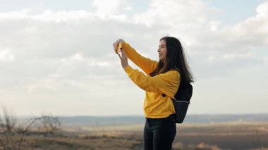 Seyahat blogcusu kızın dağın tepesinden güzel gün batımlı akıllı telefonuyla fotoğraf çekerken yan görüntüsü. Kız fotoğraf çekiyor. Tatil seyahati, mutluluk ve özgürlük yaşam tarzı kavramı