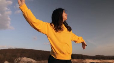 Sarı kazaklı bir kadın bir dağın tepesinde duruyor kollarını kaldırmış uçuyor, dans ediyor ve eğleniyor rüzgar saçlarını savururken - özgürlük, doğa, seyahat, günbatımı