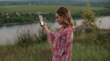 Genç ve güzel bir kadın doğada inanılmaz manzaralı bir yerde yürüyor ve akıllı telefon, blog yazarı, yürüyüş, güzel bir yer, kırsal kesimde video çekiyor.