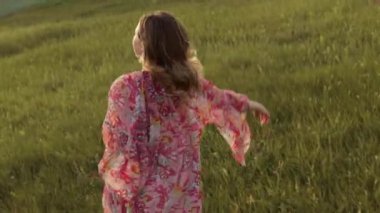 Kamera genç ve güzel bir kızı takip ediyor. Parkta çimenlerin üzerinde koşuyor. Uzun bir elbise içinde, gülümsüyor, kaygısız neşeli bir hayat, tatil, manken, eğlence anı, rahatlama.