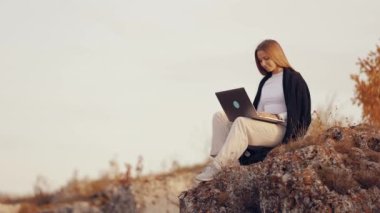 Dizüstü bilgisayarda çalışırken, doğada taş üstünde oturan serbest çalışan genç bayan öğrenci, sonbahar günü internette çalışıyor.