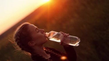 Yan görüşü kapat, genç güzel kız yürürken bir şişeden su içiyor, rüzgarlı bir sonbahar gününde güzel bir manzarayla birlikte gün batımında bir tarlada duruyor.