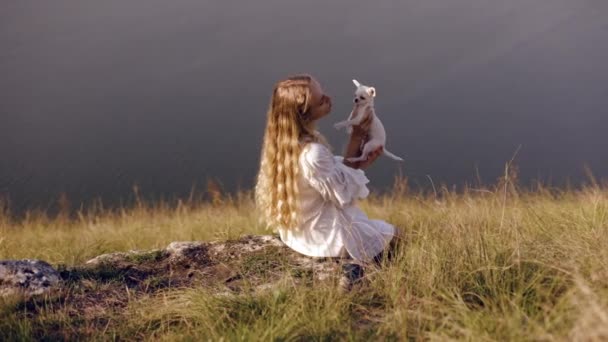 一个年轻的长发女孩坐在山坡上 带着难以置信的河景 抱着爱抚着她心爱的白雪公主犬 这是一个爱抚爱抚爱抚的概念 — 图库视频影像