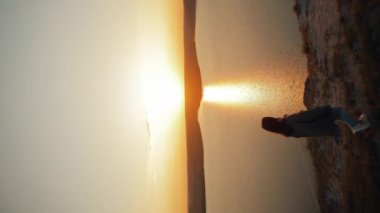 dikey video. Kamera, günbatımında inanılmaz bir manzarası olan geniş bir nehrin üzerindeki uçurumun kenarına yaklaşan ve uçma taklidi yaparak kollarını yana kaldıran bir kadın turisti takip ediyor.