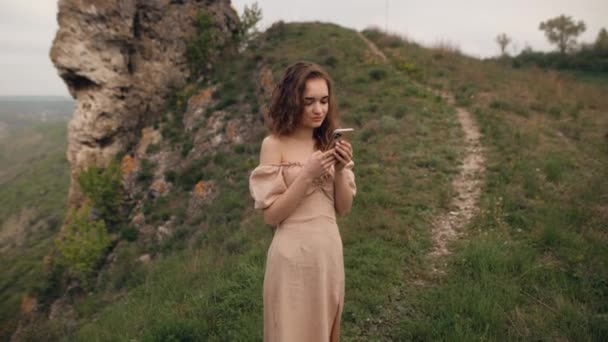 一个身材苗条的女人在旅行时使用智能手机 一个旅游姑娘站在山坡边 目光远大 手里拿着一部智能手机 — 图库视频影像
