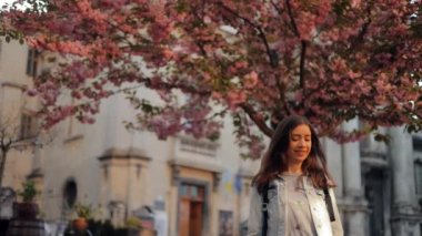 Sıradan giyimli genç bir kadın yemyeşil bahar çiçekleri olan bir sakura ağacının yanında yürüyor. Sevimli, gülümseyen bir kız Lviv şehrinde seyahat ederken yandan besleniyor.