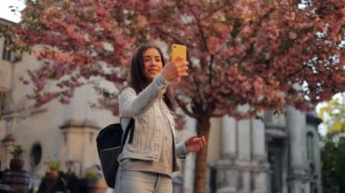 Sıradan giyimli genç bir kadın, çiçek açan bir sakura ağacının yanında dururken akıllı telefon özçekimi çeker. Şirin bir bayan seyahat ederken sosyal medya için fotoğraf çekiyor.