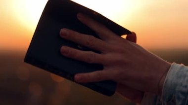 İncili okuyan, dua eden, elini İncil 'in üzerine koyan, doğada bir taşın üzerine oturan, sonbahar akşamında gün batımı, huzur ve sükunet, inanç, Tanrı' nın lütfu.