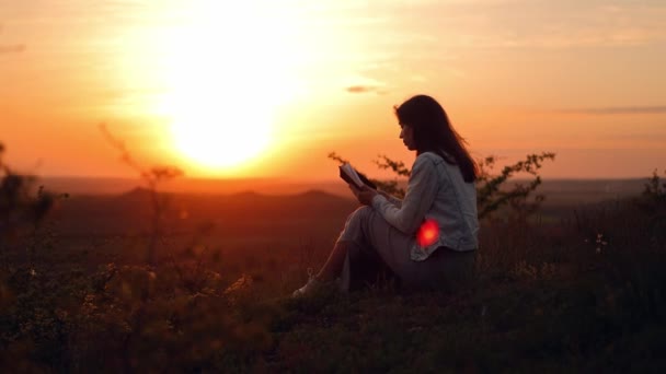 長髪の少女が太陽の下で本を読んでいる 若い女性が屋外で聖書を読みます 女性は手に聖書を持ち 山の上で神の言葉を研究しています 真実を探す — ストック動画
