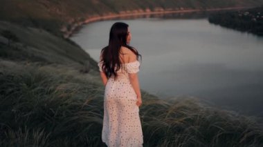 Çekici, uzun saçlı bir kadın uzun bir yaz elbisesiyle bir tepede duruyor otların arasında ve geniş nehir manzarasına hayran.