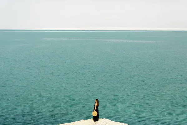 Siyah uzun elbiseli ve elinde hasır bir şapkayla genç bir kız İsrail 'de Ölü Deniz kıyısında duruyor. Panoramik çekim, yan görünüm