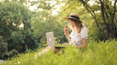 Parktaki çimenlerde oturan, dizüstü bilgisayara bakan, dondurma yiyen, lezzetli ikramların ve güzel güneşli bir günün tadını çıkaran, şapkalı çekici genç bir kadın.