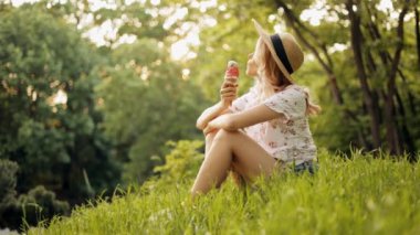 Parktaki çimenlerde oturan, dondurma yiyen, güzel ve güneşli bir tatil gününün tadını çıkaran şapkalı genç ve çekici bir kadın.