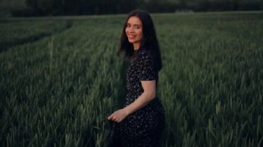 Kırsal kesimde, uzun elbiseli, yeşil buğday tarlasında yürüyen, kameraya neşeyle gülümseyen ve flört eden çekici bir kadın. Doğada yaz tatili.