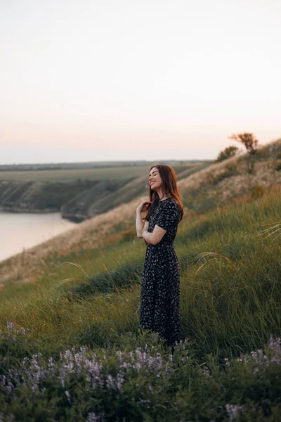 Güzel, kaygısız, güler yüzlü, gözleri kapalı bir kadın nehrin yukarısındaki uçurumun kenarında duruyor güneş batarken kır çiçeklerinin arasında, doğanın ve yaz tatilinin tadını çıkarıyor.