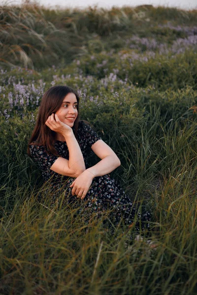 Uzun elbiseli bir kadın gün batımında elinde mor çiçeklerle bir çayırda oturur. Yaz manzarası