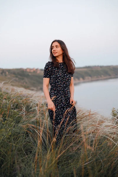 Uzun siyah saçlı, güzel, beyaz bir kadın. Uzağa bakıyor ve gülümsüyor. Denizin yukarısındaki volkanik bir kayada poz veren uzun elbiseli şirin bir kadın portresi.