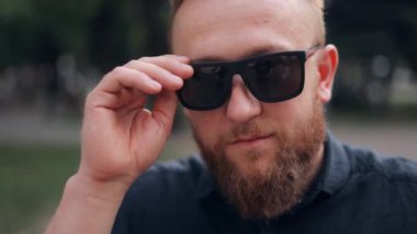 Genç sakallı bir adamın yakın plan portresi. Gözlüğünü çıkarıyor ve gülümseyen kameraya bakıyor.