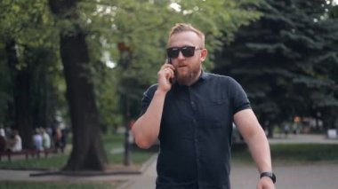Güneş gözlüklü, genç, sakallı, beyaz iş adamının şehirde yürüdüğü ve iş ortağıyla cep telefonundan konuştuğu yakın plan..