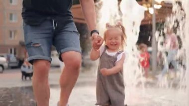 Bir baba, gün batımında fıskiyeden akan suyun içinde onunla birlikte parkta koşan küçük kızının elini tutar. Aile mutluluğu. Çocuk çok mutlu ve neşeli. Güzel hayat.
