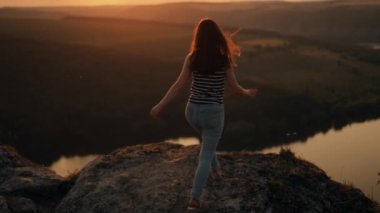 Gün batımında dağlarda büyük bir taşın üzerinde tek başına duran bir kadın yürüyüşçünün silueti. Kadın turist, akşam vakti elini yüksek kayalara kaldırıyor. Turizm, seyahat ve sağlıklı yaşam tarzı konsepti.