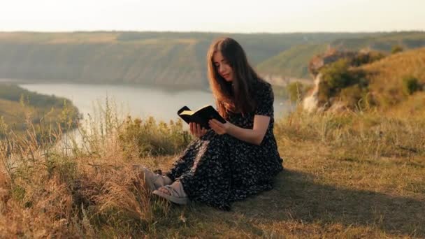 美丽的年轻长发女子坐在草地上 有着不可思议的景色 手里拿着一本圣经 阅读着 仰望着风景 欣赏着美丽 愉快地闭上眼睛 — 图库视频影像