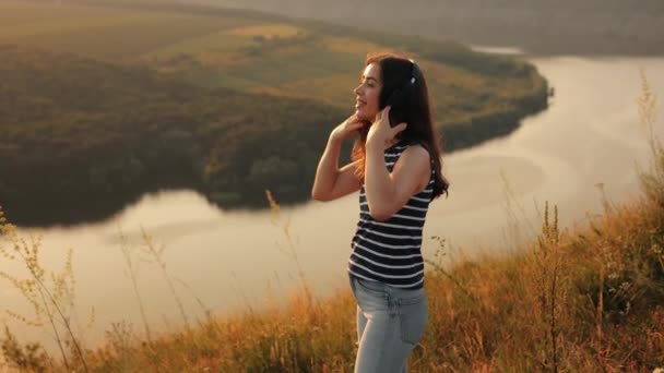 一个年轻姑娘 背景是河流和高山的本质 夕阳西下时 喜欢用耳机听音乐 一边唱着歌 一边欢快地跳舞 一边欣赏着摄像机 — 图库视频影像