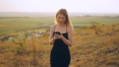 Siyah elbiseli güzel, sıska bir kadın elinde akıllı bir telefonla yürüyor. Gün batımında bahçedeki çimlere bakıyor ve büyüleyici bir şekilde gülümsüyor.
