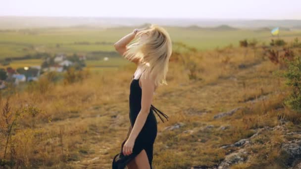 穿着黑色衣服的美丽纤细的年轻女子一边走着 一边看着夕阳西下在草地上盘旋的相机 迷人地微笑着 调情着 — 图库视频影像
